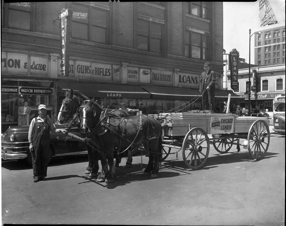 Horse-drawn carts and a parked car on a busy city street in front of storefronts.