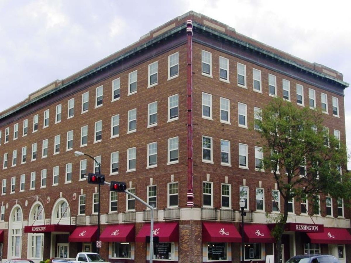 Corner brick building with red awnings and storefronts on the ground floor at a city intersection.
