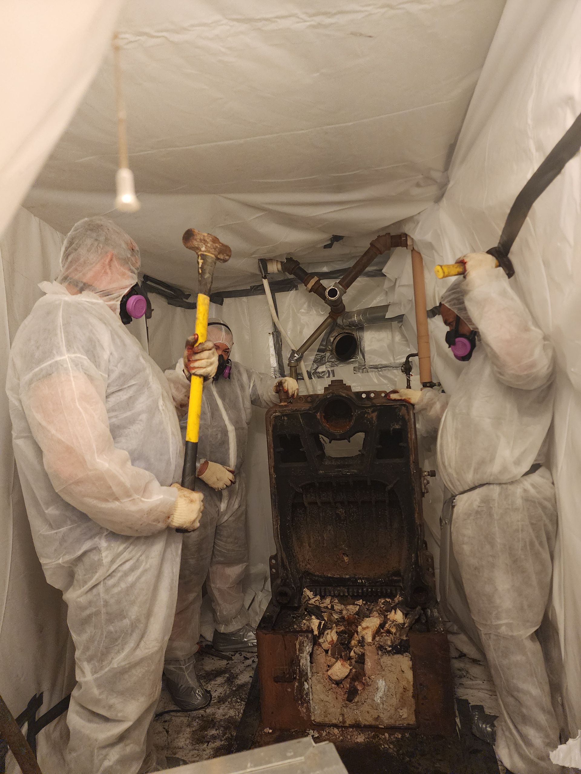 Three people in protective suits removing a furnace, working inside a white enclosure.