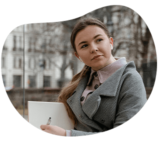 Woman wearing headphones and a gray coat, holding a laptop, looking off to the side. Outdoor setting.