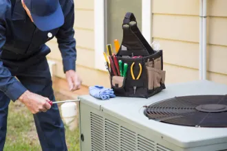 HVAC technician in blue overalls servicing an air conditioning unit outside, with a tool bag.