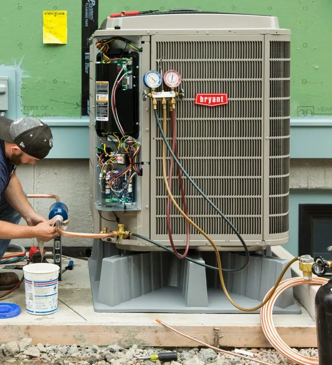 HVAC technician repairing a Bryant AC unit; using gauges and copper tubing outdoors on a concrete pad.