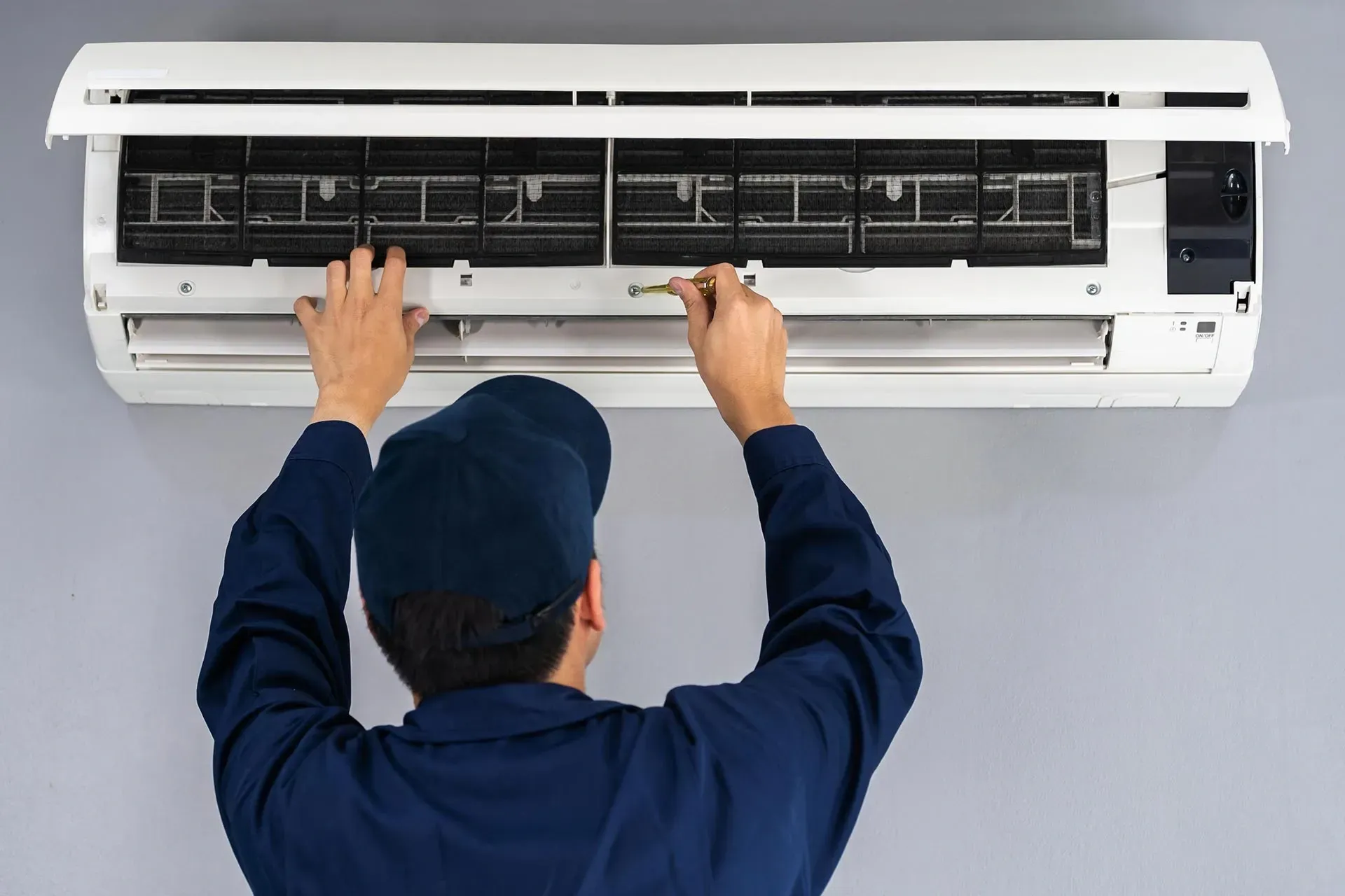 Person in blue uniform cleaning a wall-mounted air conditioning unit; hands are visible, gray wall background.