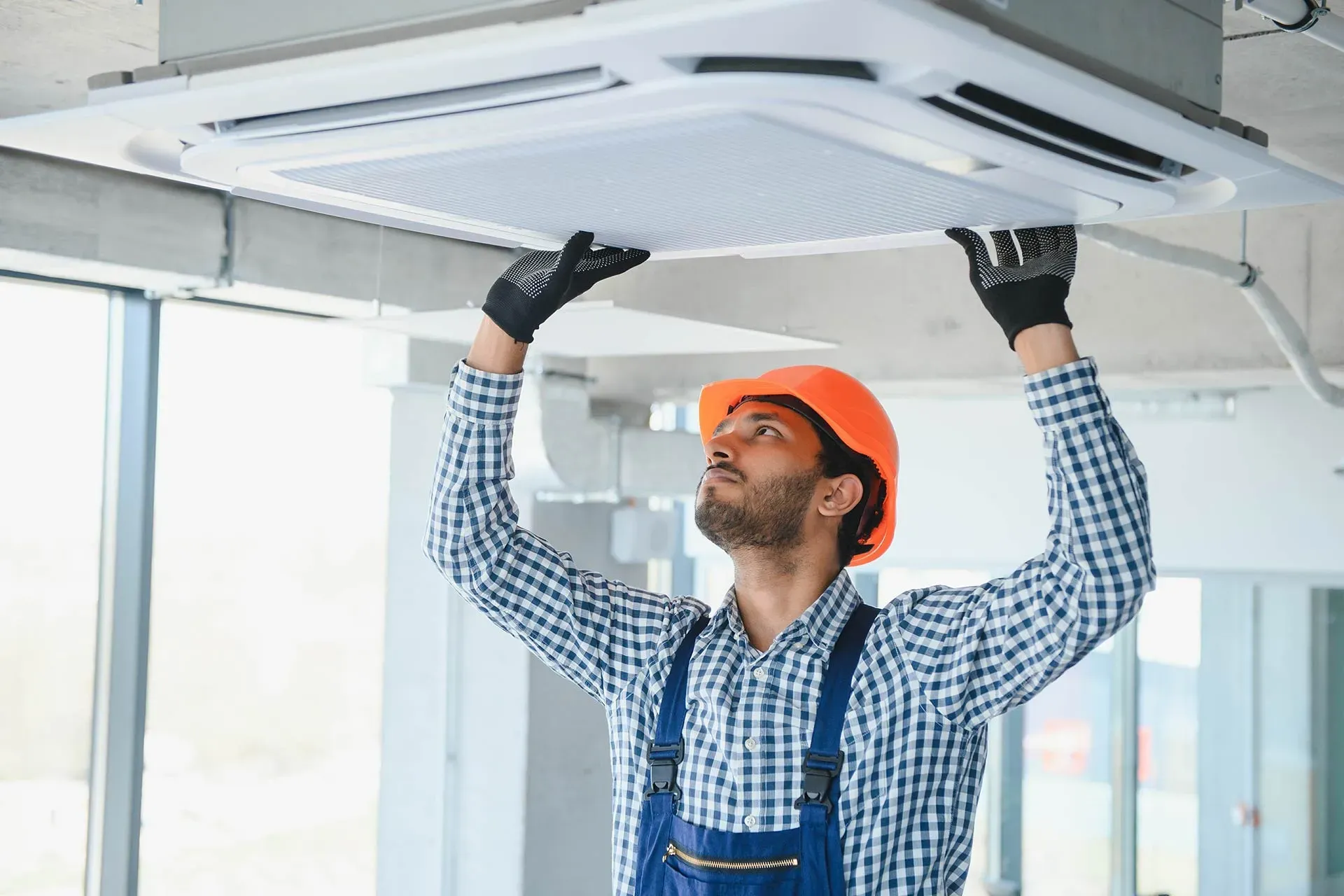 Man in overalls and hard hat installing ceiling air conditioning unit.