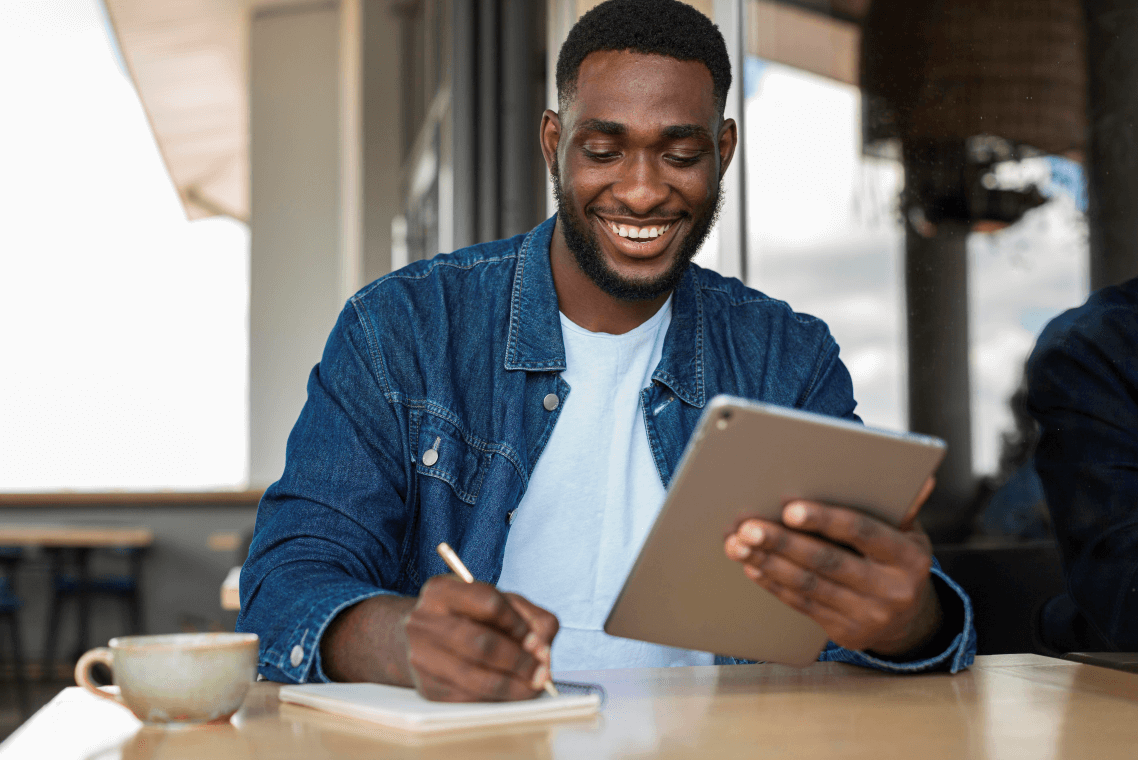 A man is sitting at a table using a tablet and writing in a notebook.