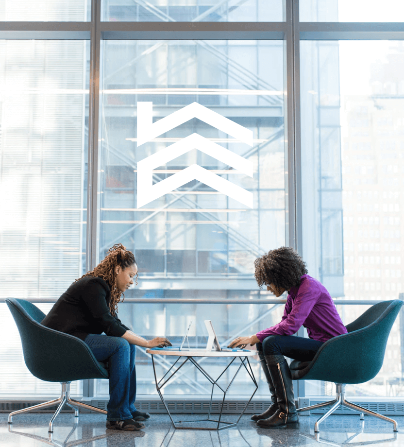 Two women are sitting at a table with laptops in front of a window.