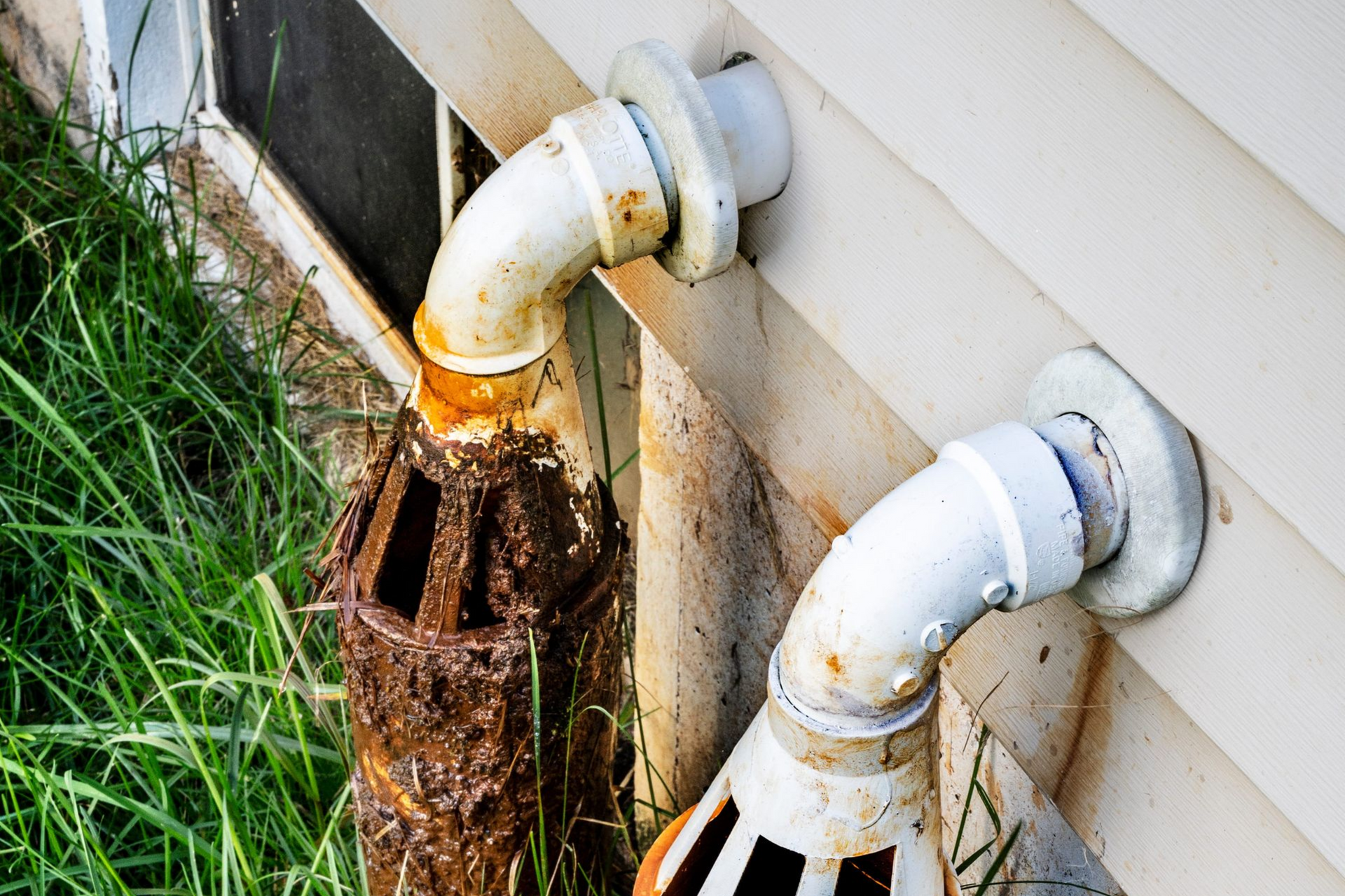Rusty pipes exiting a building's siding near a window. Grass surrounds the pipes, which have white flanges.