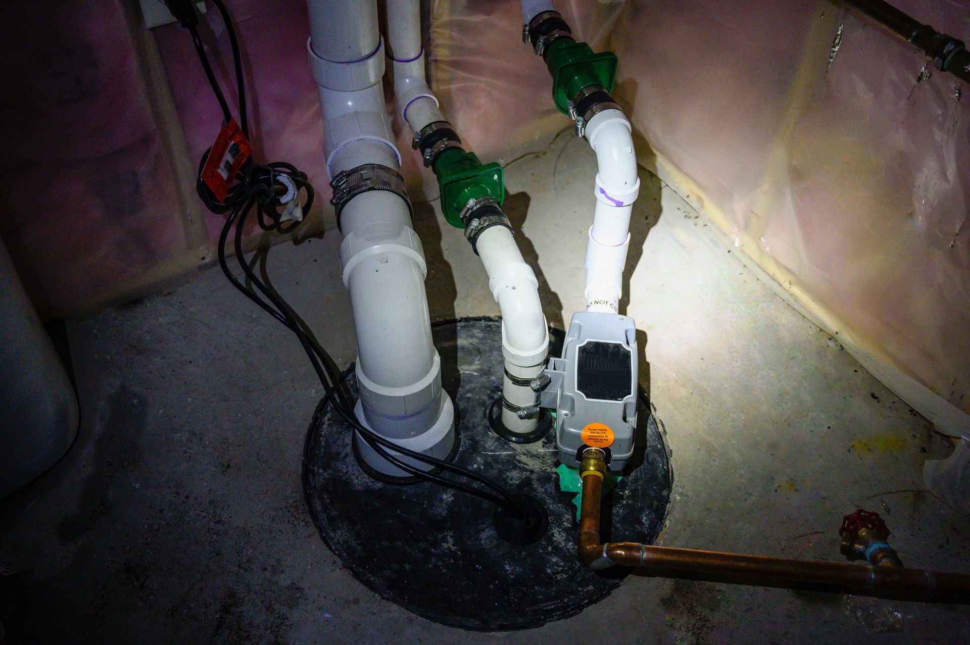 Sump pump system with white PVC pipes, green valves, and electrical wiring in a basement setting.