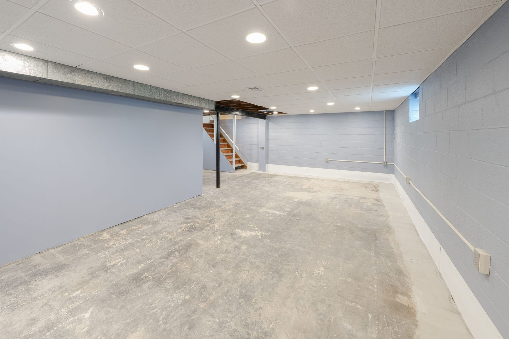 Empty basement with gray concrete floor, light blue walls, and recessed lighting. Staircase visible.