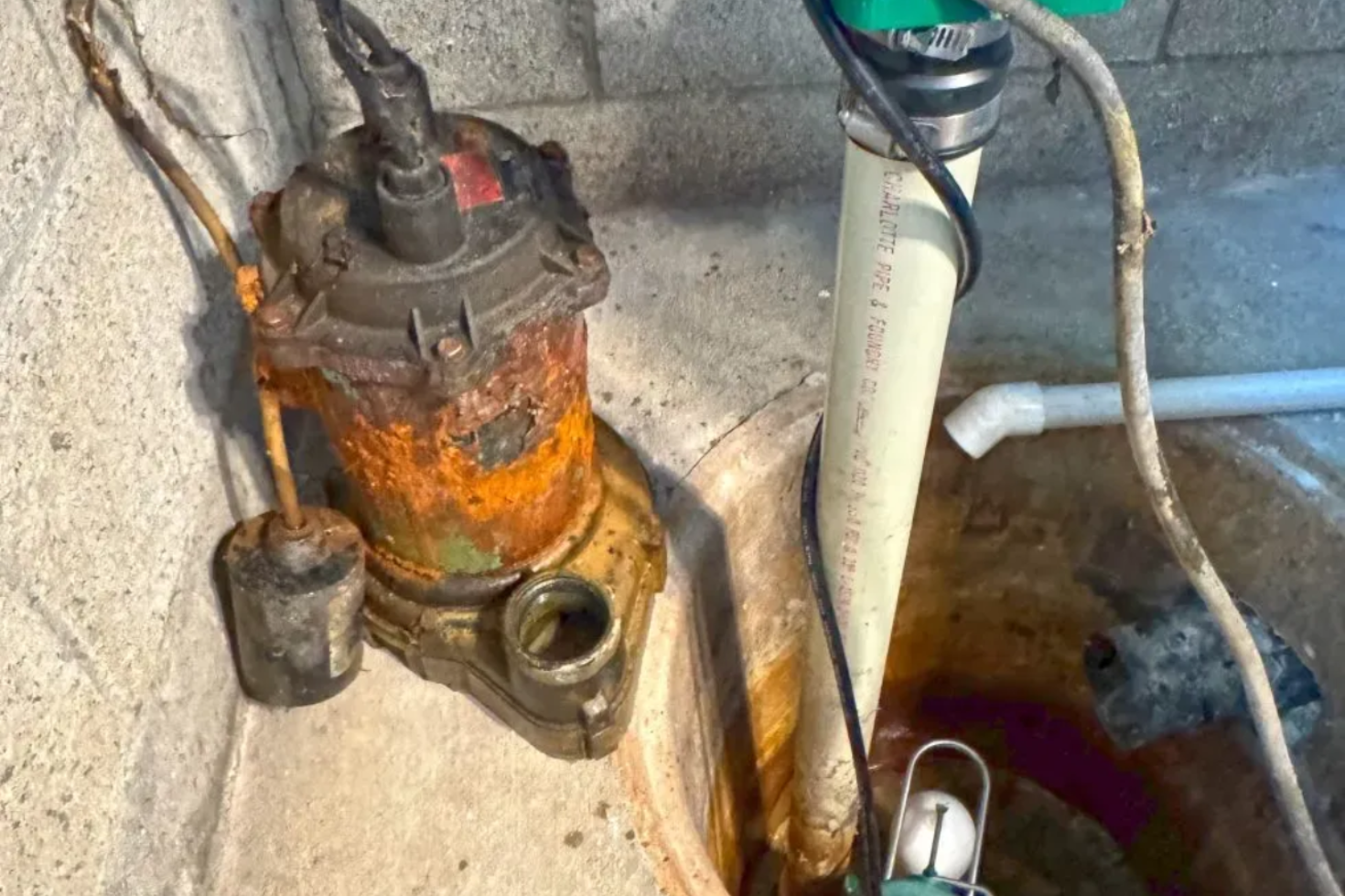 Rusty sump pump in a concrete pit with PVC pipe and electrical cords.