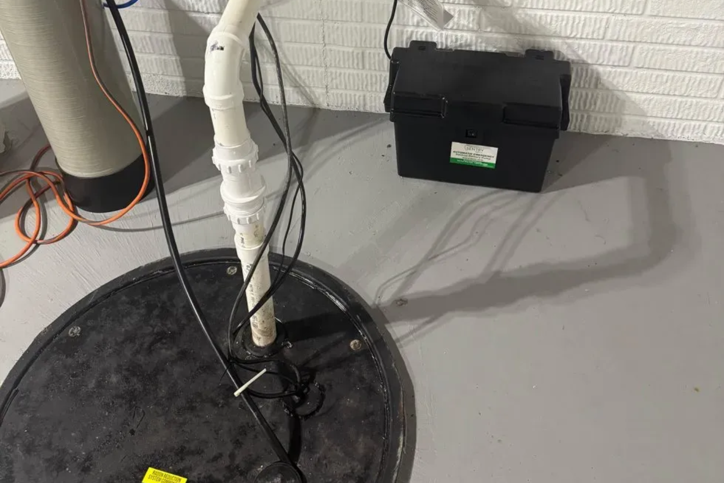 A sump pump system in a basement with a battery backup. Black round basin, white PVC pipe, black battery box.