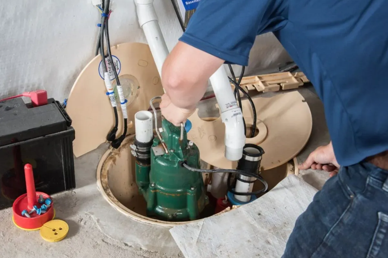 Man working on a sump pump in a basement. The pump is green, and the man is wearing a blue shirt.