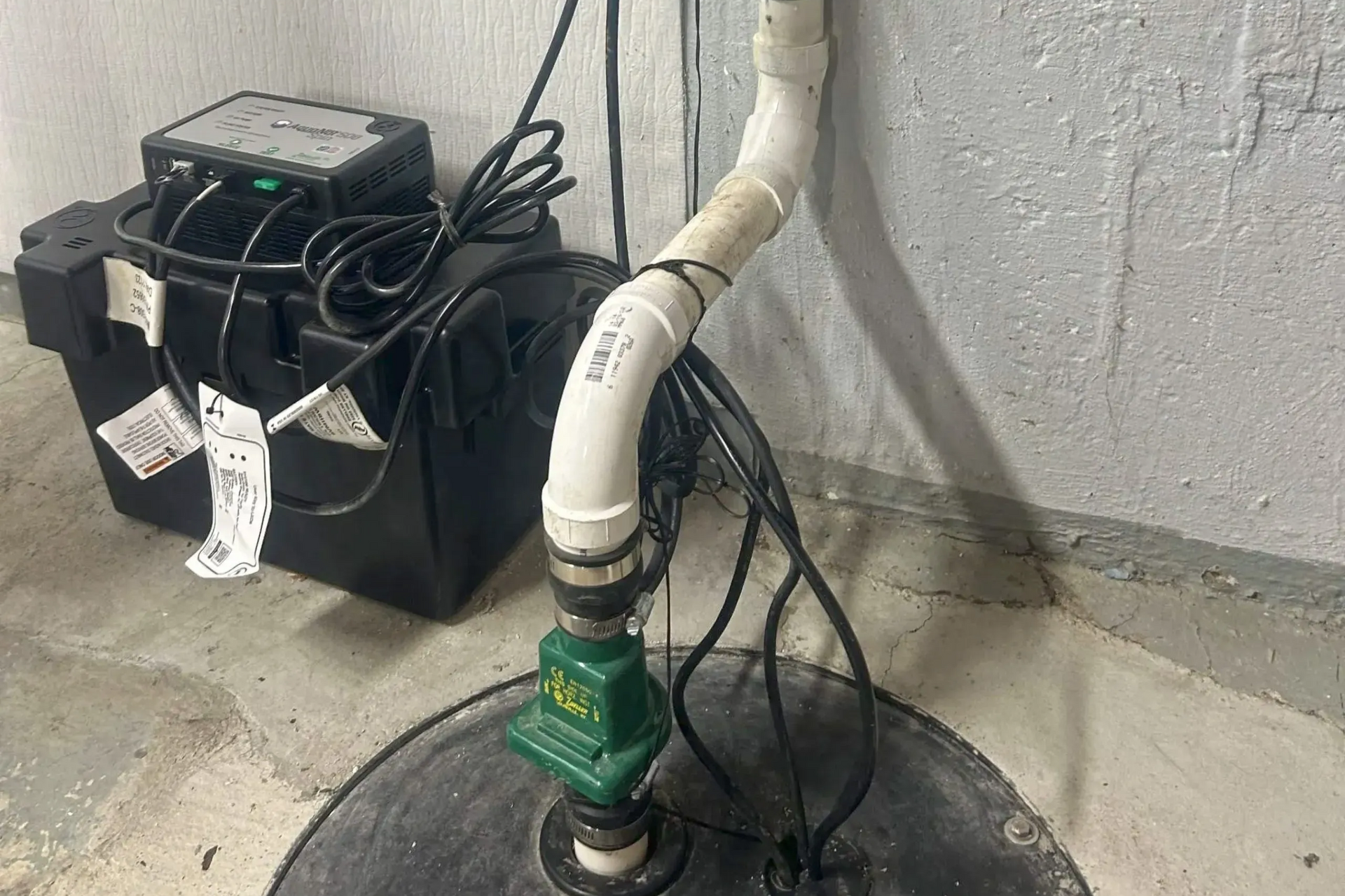 Sump pump system with black pump, battery backup, and white PVC pipe in a concrete basement.