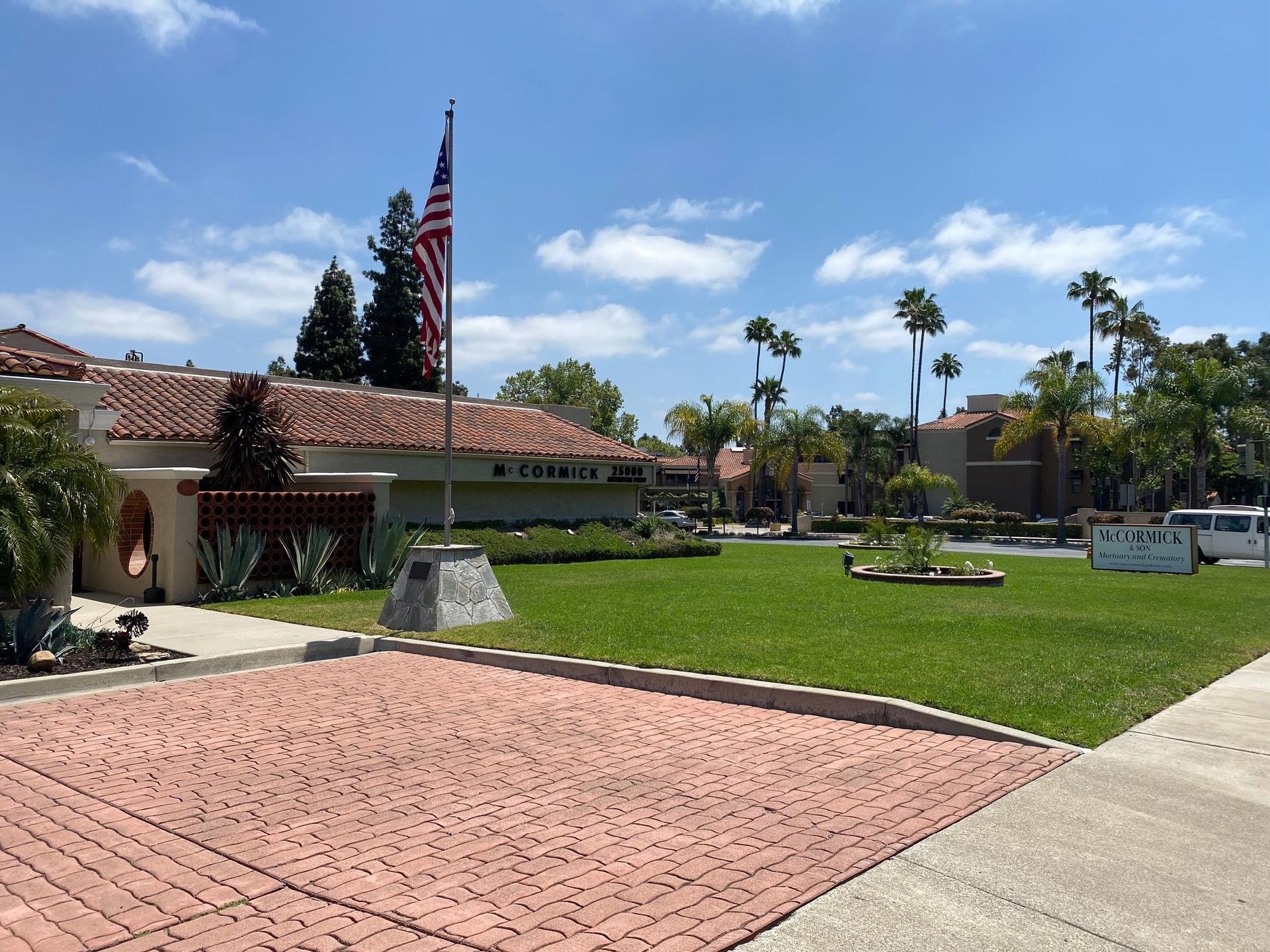 American flag waving in front of a building with red tile roof and a brick-paved area on a sunny day.