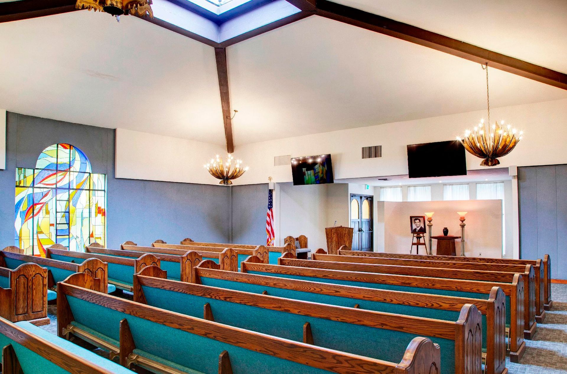 Rows of wooden pews in a funeral home chapel, with a stained-glass window, two chandeliers, and an American flag.