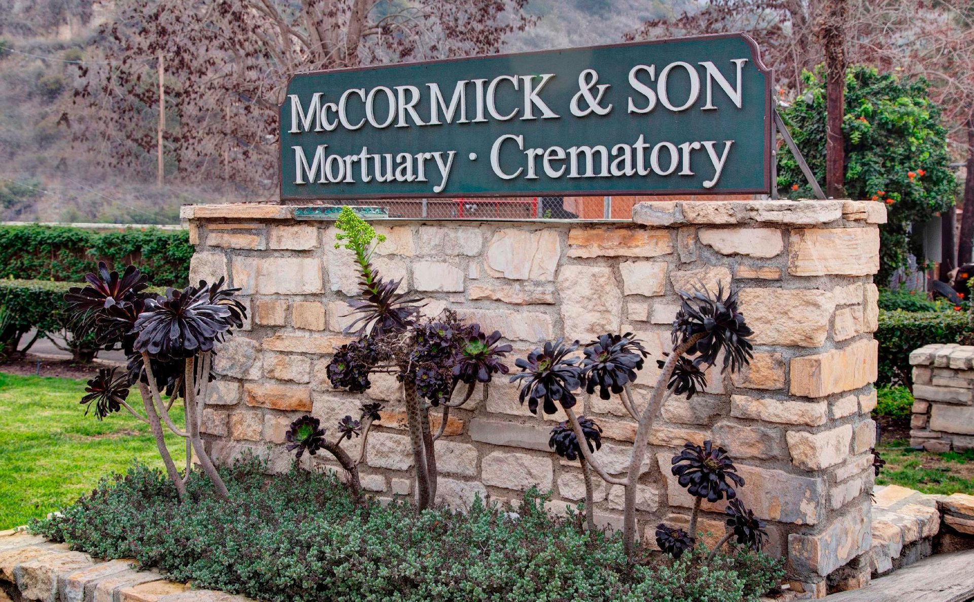 Sign for McCormick & Son Mortuary and Crematory on a stone wall with dark plants in front.