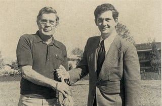Two men shaking hands outdoors; one in a polo, the other in a suit.