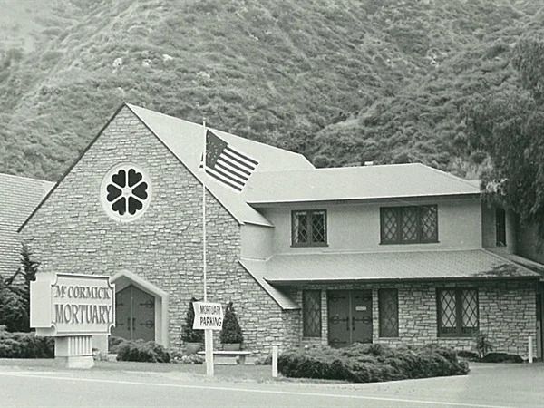 Mortuary with stone facade, flower window, sign, flag, and mountain backdrop.
