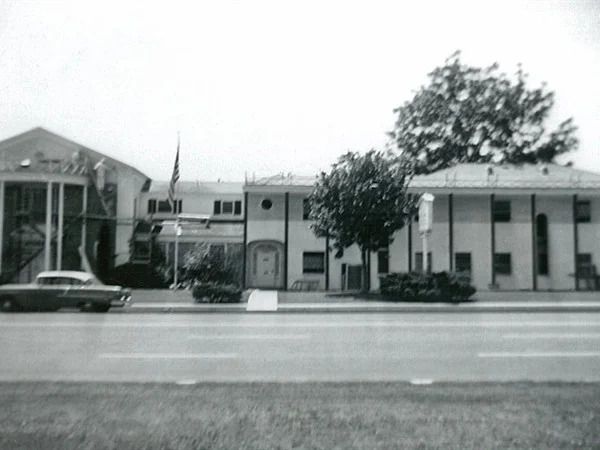 Black and white photo of a building with columns, an American flag, and a car parked on the side.