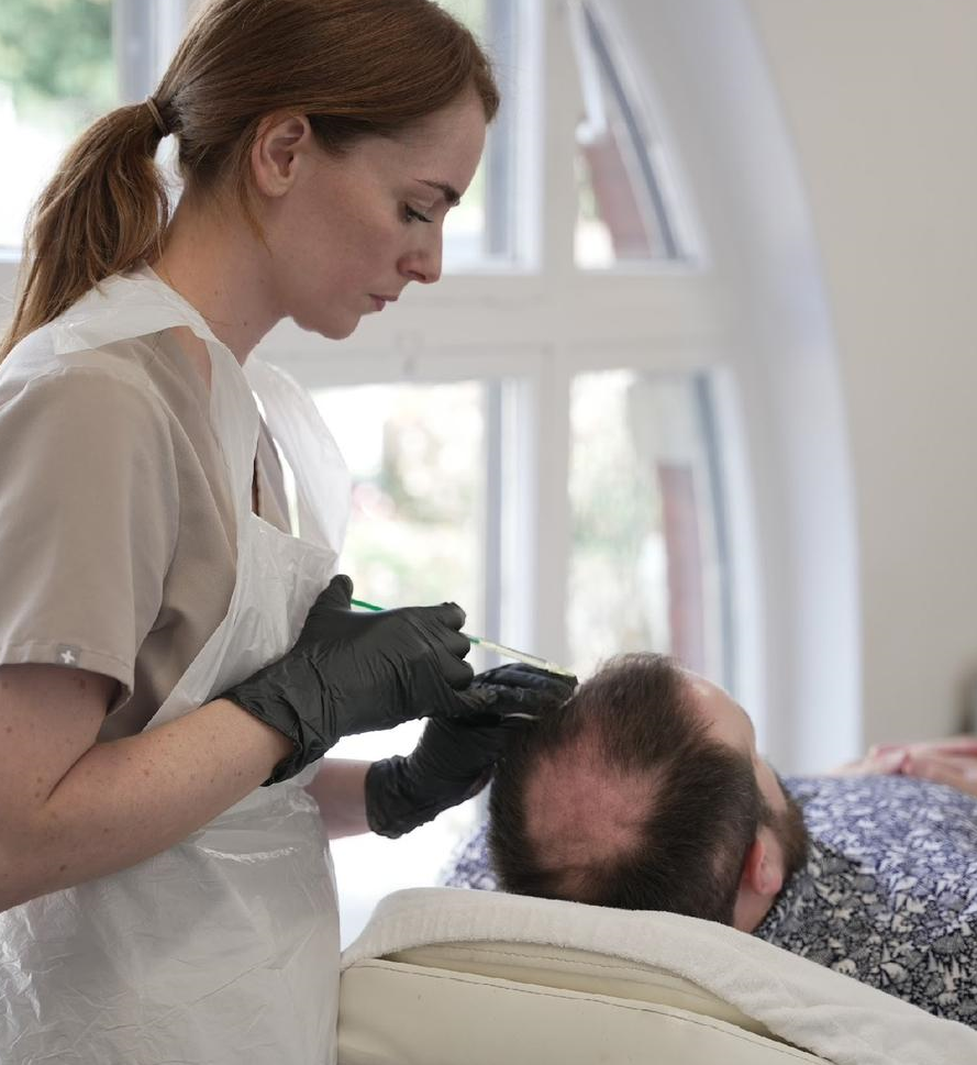 Woman administering scalp treatment to a man in a clinic, wearing gloves and apron.
