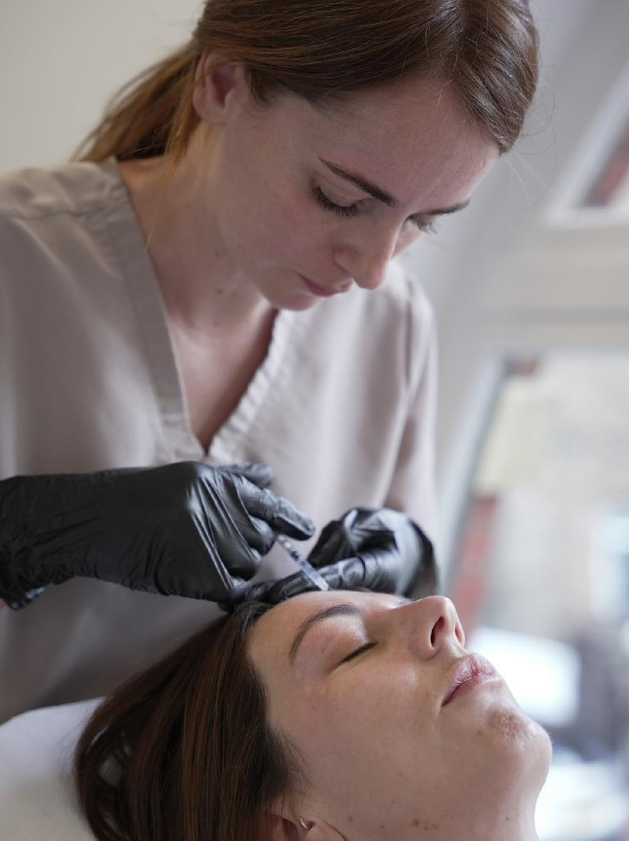 Beautician administering an injection to a client's forehead. Dark gloves and neutral-toned scrubs.