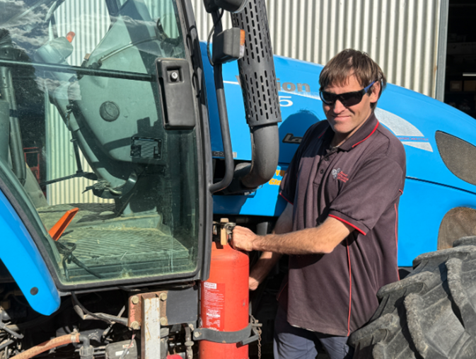 A man in a polo shirt and sunglasses standing next to a blue tractor, placing a red fire extinguisher into a mounting bracket.