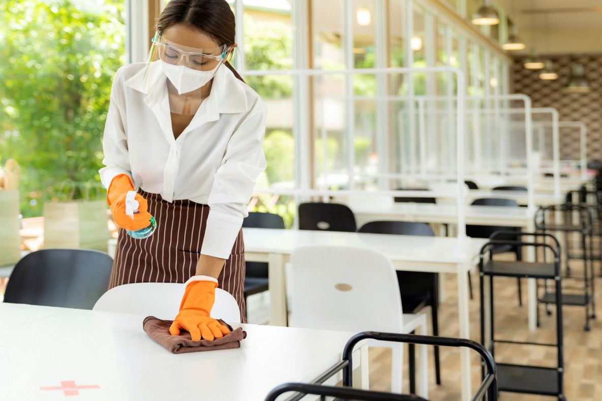 A woman wearing a mask and gloves is cleaning tables and chairs in a restaurant.