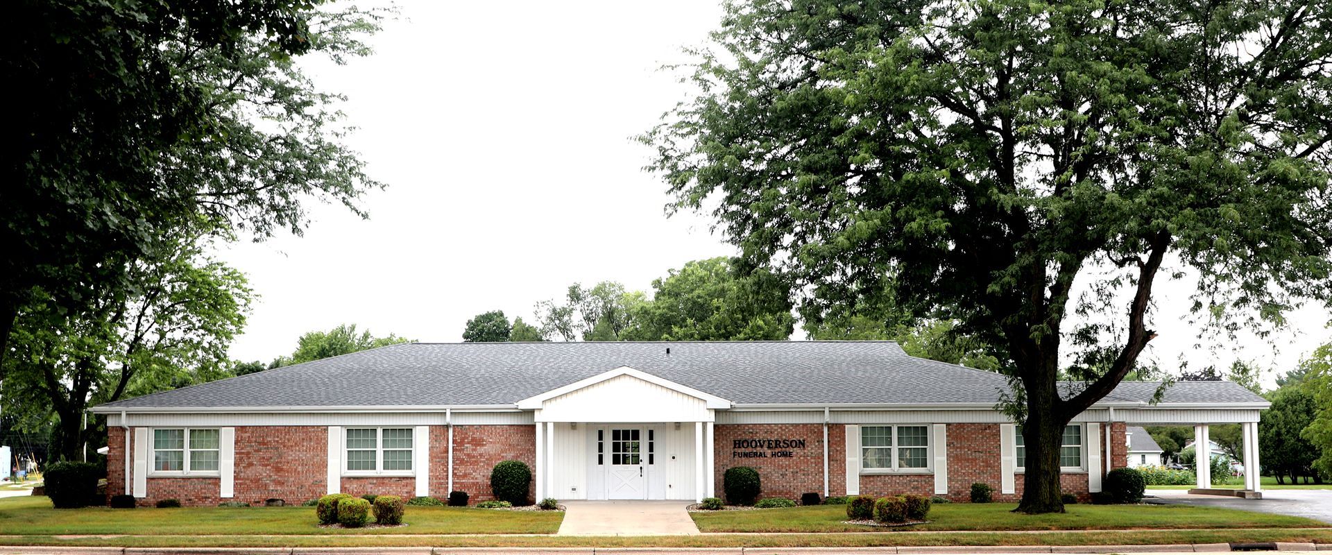 A brick building with a dark roof and white trim, flanked by large trees.
