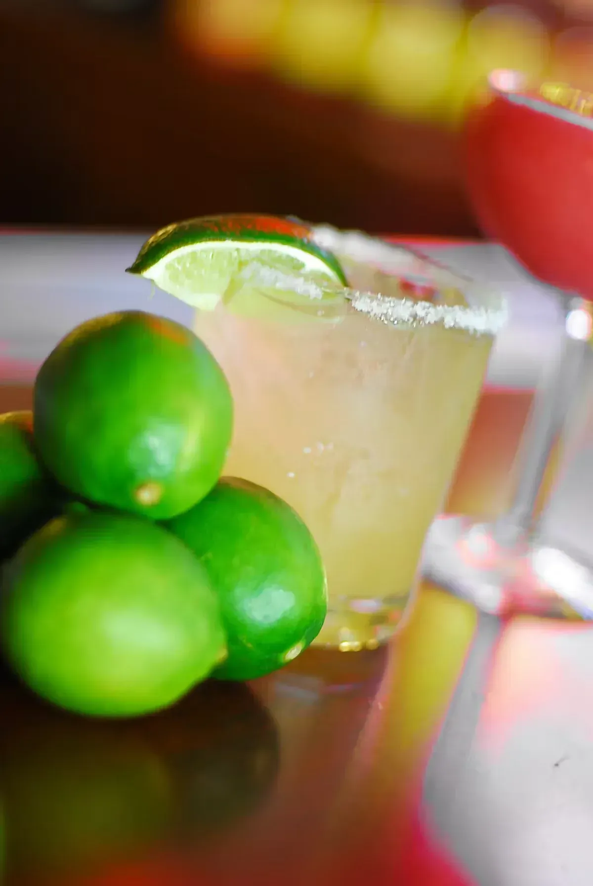 Margarita with lime wedge garnish and salt rim, limes stacked nearby, on a reflective surface.