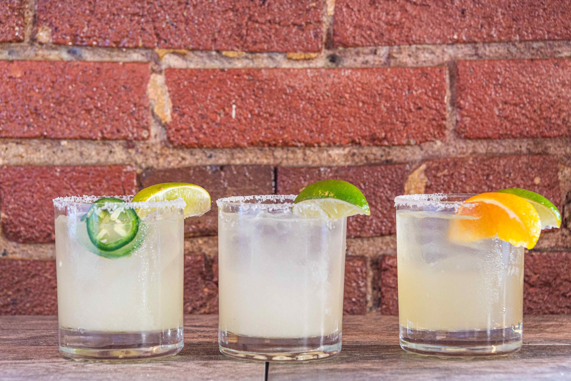 Three margaritas with salt rims, lime and orange wedges, and jalapeño on a wood surface, against a brick wall.