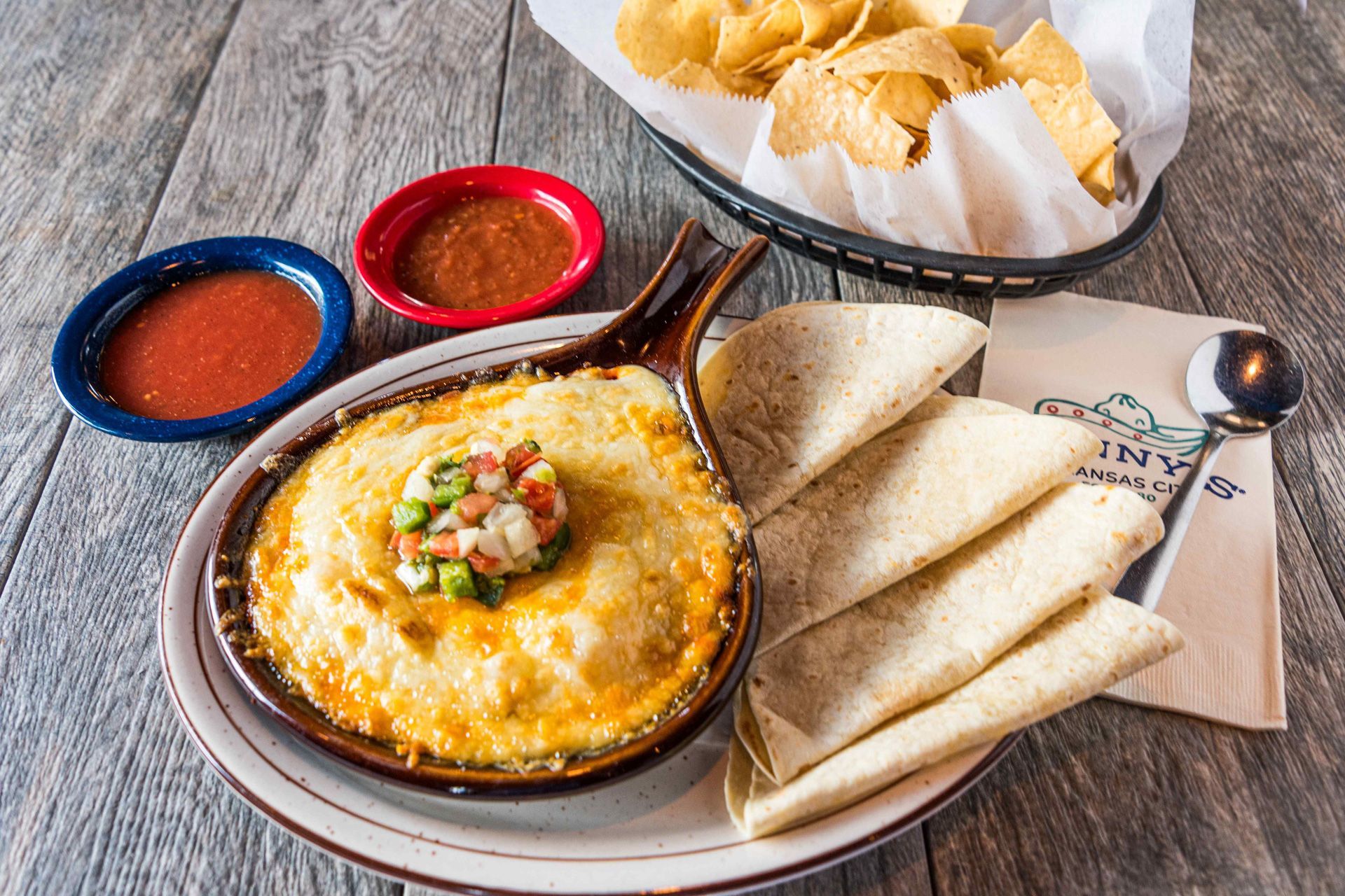 Baked dip with toppings, salsa, tortillas, and chips in a basket on a wood table.