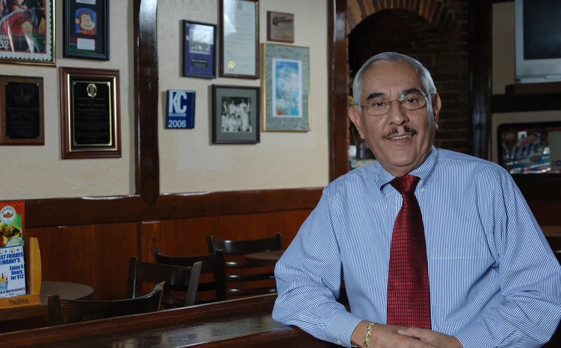 Man in blue striped shirt, red tie, smiling, leaning on bar; framed items on wall behind.