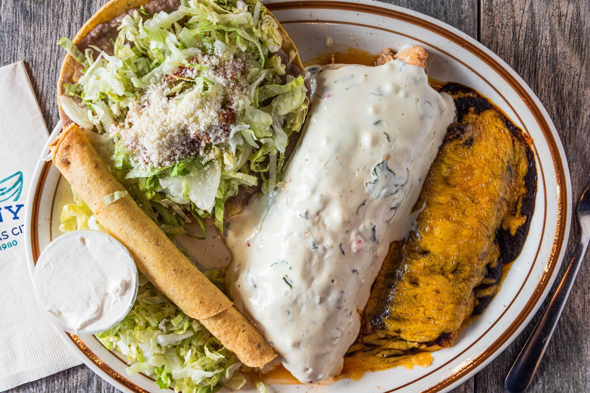 Plate with a smothered burrito, tostada, taquito, and sour cream.