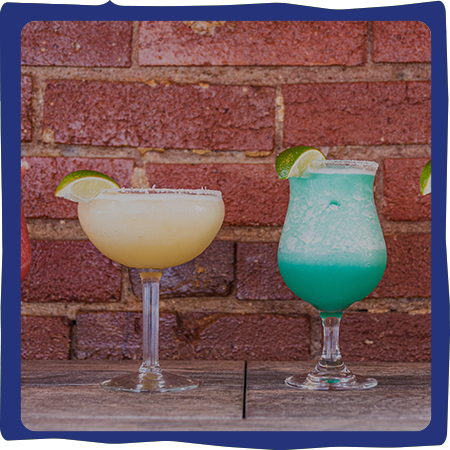 Two margaritas with lime garnishes, one yellow, one blue-green, sit on a table in front of a brick wall.
