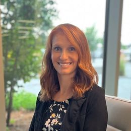 A woman with red hair is sitting in front of a window and smiling.