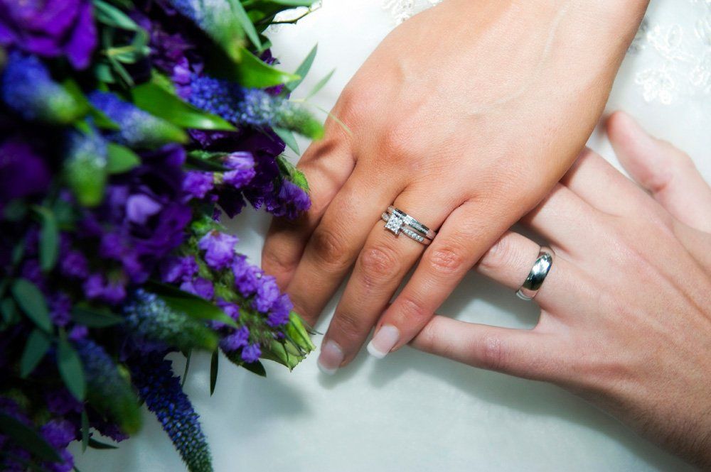 Wedding rings on joined hands with a purple flower bouquet.