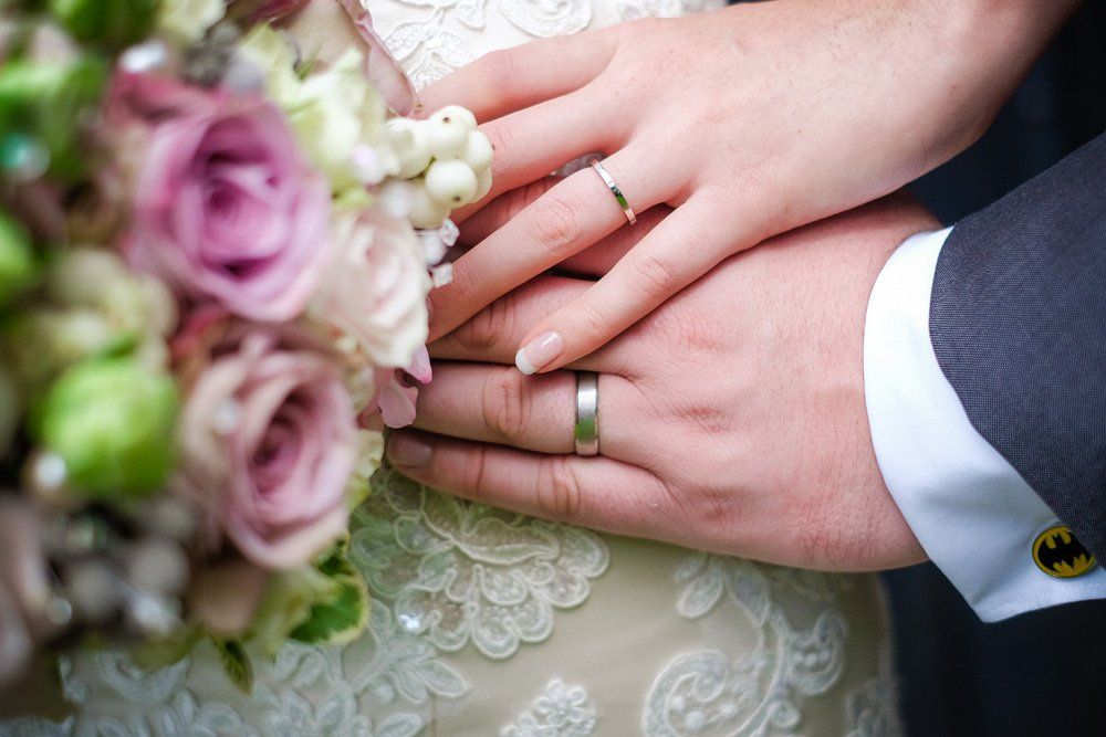 Wedding rings on clasped hands with bouquet and lace dress.