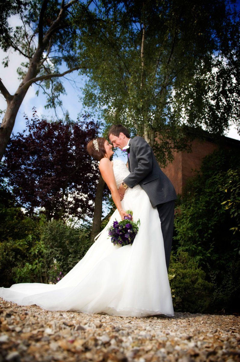 Bride and groom embrace outdoors, she in a white gown, he in a gray suit; trees and a building in the background.