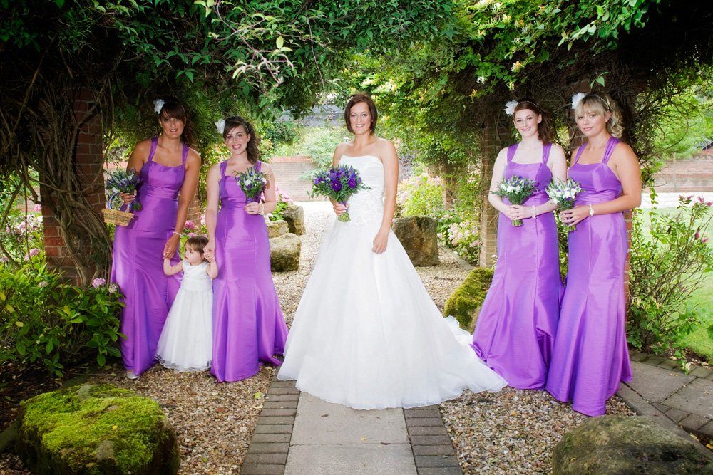 Bride in white gown with bridesmaids in purple dresses, garden setting.