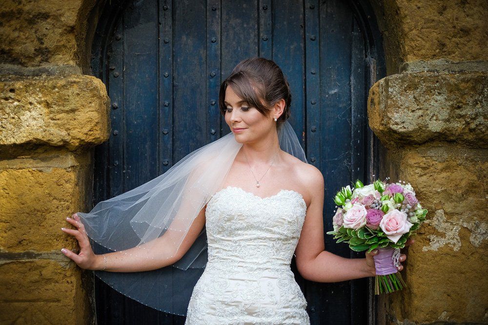 Bride in white strapless dress, holding flowers, against a blue door. She wears a veil and smiles.