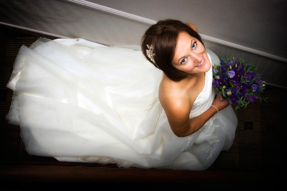 Bride in a white gown holding purple flowers, looking up and smiling on a staircase.