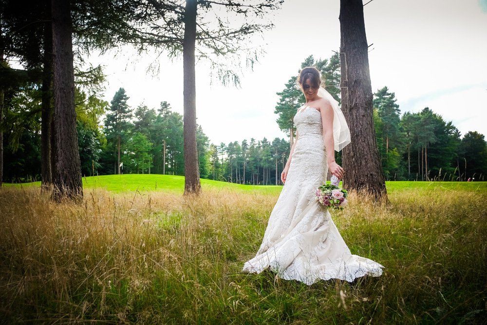 Bride in a white gown holding flowers, stands in tall grass among trees on a sunny day.