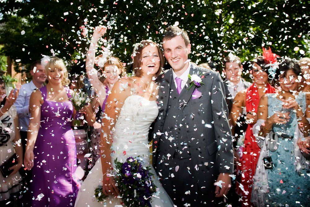 Newlyweds showered with confetti, surrounded by wedding guests celebrating outdoors.