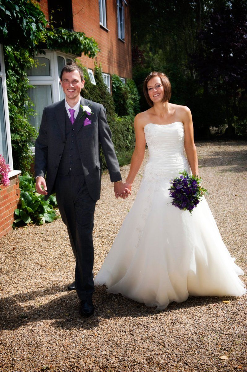 Bride and groom walking, holding hands. Bride in white gown, groom in suit, outside brick building.