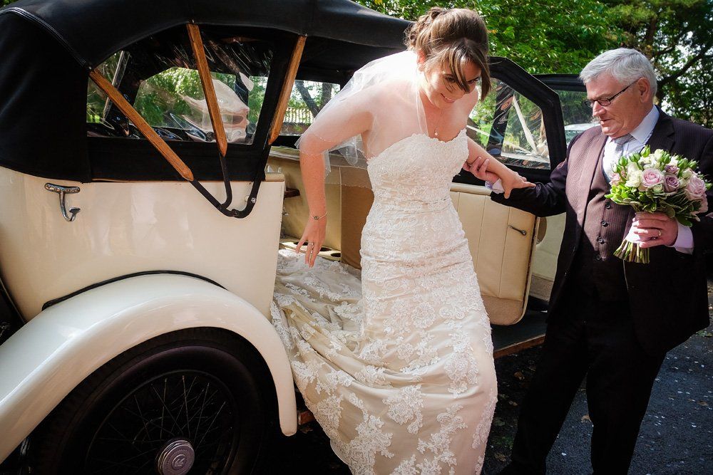 Bride exiting classic car, assisted by a man holding flowers. White dress, beige car, outdoor setting.
