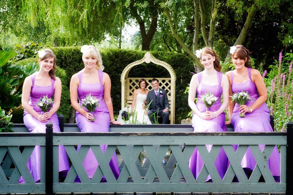 Bridesmaids in purple dresses holding bouquets, bride and groom in background under an arbor.