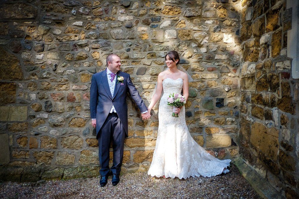 Bride and groom hold hands, smiling, in front of a stone wall. He wears a suit; she wears a wedding dress.