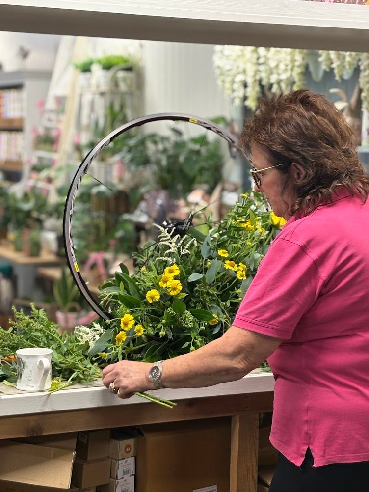 Woman arranging flowers in a bicycle wheel, shop setting with plants.