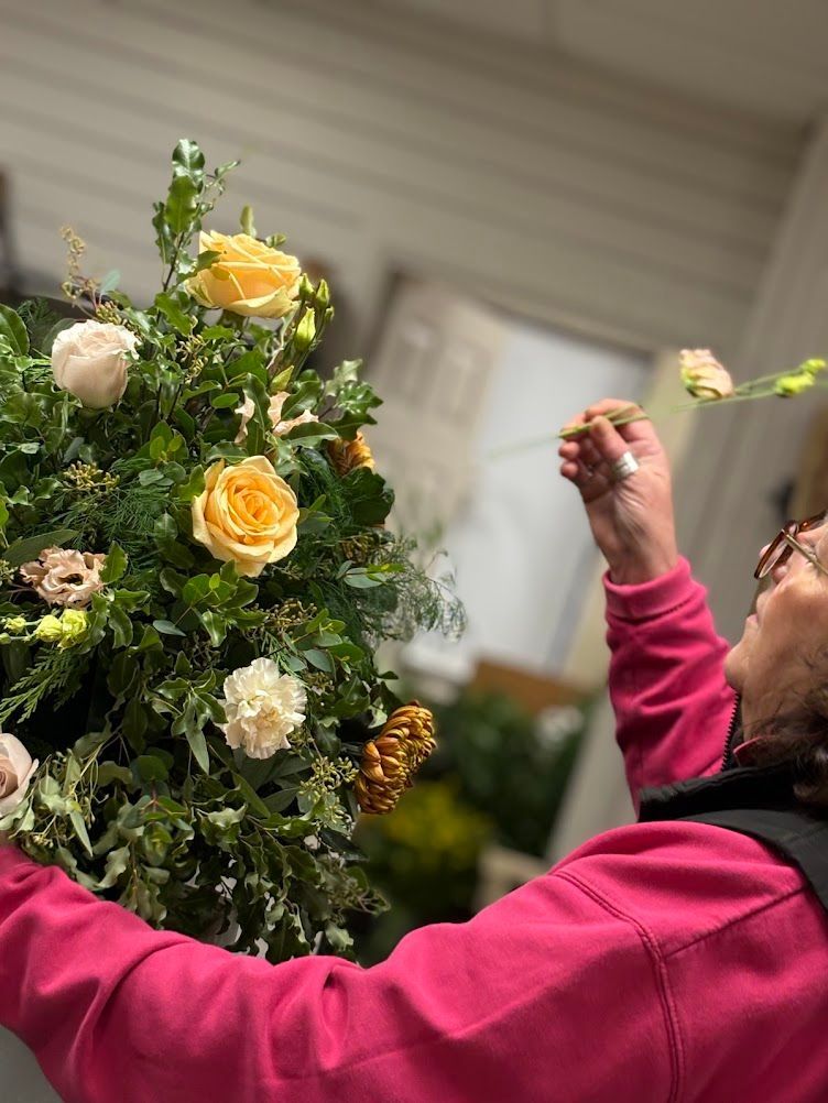 Person arranging flowers, wearing pink and black. Yellow roses and cream flowers in the arrangement. Interior setting.