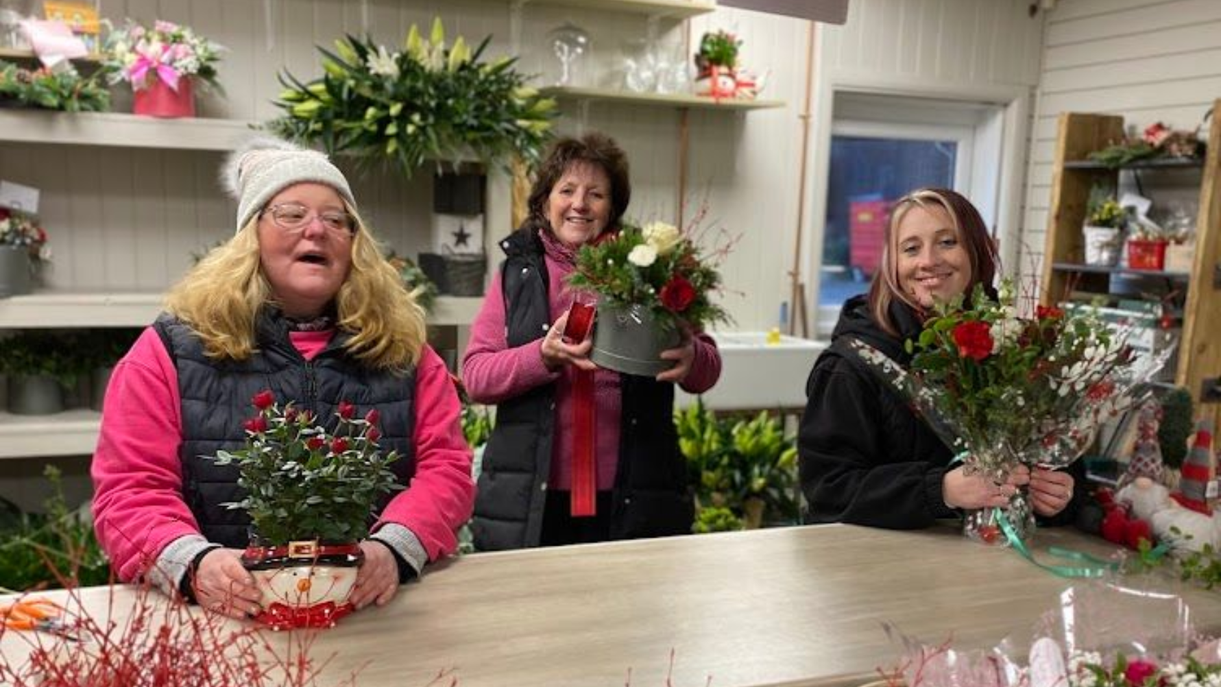 Three women in a flower shop, holding floral arrangements. One wears a winter hat and vest.